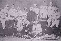 A group of men wearing their team uniforms, some standing and some sitting, each with a hockey stick, in a studio. On an easel is an Ottawa championship banner. On a table is a trophy.