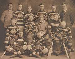 Thirteen men are assembled in three rows, two sitting on the floor in front, five sitting and six standing in back. Ten are hockey players in their uniforms with their hockey sticks and three are members of the team staff in suits.
