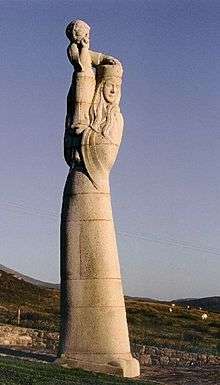  A large stone statue of a tall and slender woman by a field. The woman has long hair and wears a hat and carries a child on her shoulders.
