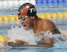 Brown skinned man wearing goggles and a cap with the black and red colours of his country and the flag of Tunisia, wearing a swimsuit not covering his torso, does breaststroke, with his arms parallel to the surface of the water. He is opening his mouth to take in a breath as his head is above the water.