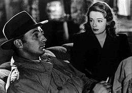 Black-and-white image of a man and a woman sitting side by side on a couch, viewed at an angle. The man, in profile in the left foreground, stares off to the right of frame. He wears a trenchcoat, and his face is shadowed by a fedora. He holds a cigarette in his left hand. The woman, to the right and rear, stares at him. She wears a dark dress and lipstick of a deeply saturated hue.