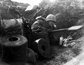A Caucasian soldier wearing a camouflage helmet and thick winter clothing lies on his side in a trench facing the right of the photo, holding a canteen in his left hand and using an entrenching tool with his right hand. The soldier is surrounded by rolls of communication line, jerries and boxes of equipment. Behind him, the skyline can be seen above the top of the earth works.