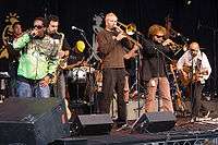 In the left a black man with sunglasses and a green jacket holding performing to a microphone beside him another man playing the saxophone, in the center blonde haired man playing the trombone and in the back a man with sunglasses and a yellow hat playing the percussion with his head down, and in the right a man with curly hair with sunglasses playing the trumpet beside him a bald man playing the guitar.