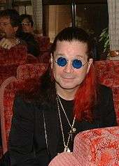 A man with red coloring on his dark hair, wearing sunglasses, necklaces, and a black suit. He is seated on a red chair, and two people are also seated in the background.