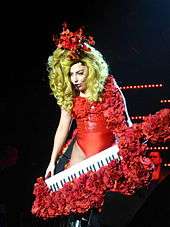 With long blonde hair, a woman holds an instrument wearing a red outfit.