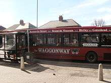 In a sunlit suburban street, a single-decker red bus is parked at a bus stop facing left. Above its windows are the words "Waggonway", "Chester-le-Street", "Beamish Museum", "Birtley" and "Gateshead". On a large window on the right is the word "Beamish", and below the windows are the words "the Waggonway" and a stylised picture of an early railway cart.
