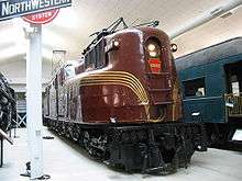 A burgundy locomotive, with gold stripes in a museum with other railroad equipment.