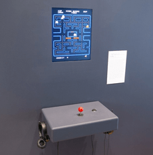 A screen on a plain gray wall displaying Pac-Man, with a simple joystick and pair of headphones beneath it