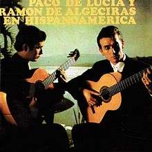 Two men playing flamenco guitar in front of a greenish tinted background depicting a body of water and mountain in the distance.