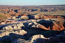 An eroded multi-colored landscape of rounded buttes and small arroyos stretches into the distance under a blank sky.