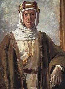 A man in white Bedouin-style robes and headdress, with a brown outer gown, from beneath which the handle of a sword or similar item protrudes