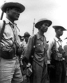 Three men, wearing uniforms and hats are standing and looking to the right. They are armed with grenades, guns, and have pouches. Other men can be seen in the background.