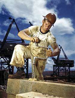 An American carpenter wearing overalls, boots, and a hard hat bores a hole with a hand drill.