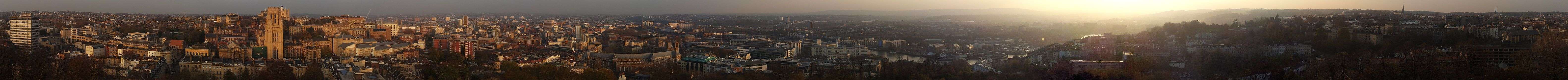 A photographic panorama of Bristol taken from the top of the Cabot Tower. The picture shows an urban environment with densely packed offices and older buildings. Hills can be seen in the distance.