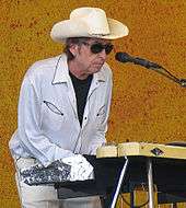 A man behind a keyboard and microphone stand, wearing a cowboy hat, sunglasses, a white dress shirt, and white pants.
