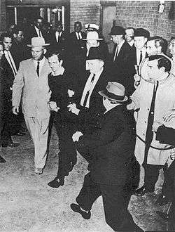 A black-and-white photograph showing a man with men on either side of him walking past people on either side, from above. At the bottom right a man with a hat has a gun pointed at the man in the center.