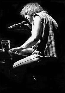 Black-and-white photo of a man playing the piano, there is a drink on the piano.