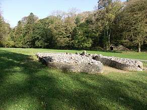 Front view of a cairn, from its right side, its boulders retained by a short wall that forms a courtyard at its entrance. The cromlech is set in flat ground of short grass (in dappled sunlight in the foreground and full sun elsewhere), dissected by a path passing behind it. Trees are mainly in leaf to its rear, among which a limestone kiln is visible at the foot of a gorge.
