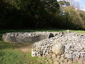 A short dry-stone wall retains boulders to form a cairn. The wall is missing at the front, right section, where the rubble has tumbled out, leaving a (previously covered) orthostat exposed. The wall forms a courtyard at the tumulus' entrance. Flat ground of short grass surrounds the cairn. The background is of shaded trees, mainly in leaf.
