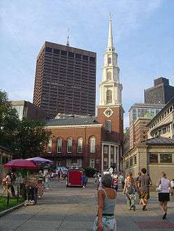 Ground level view of a brick church with a prominent white steeple; a grassy park is visible in the foreground, and a rectangular skyscraper with a tan facade and black windows is in the distance