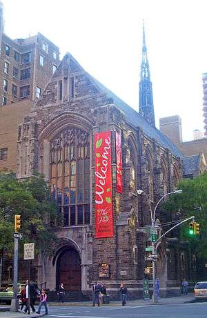 A tall brown stone church with a pointed roof and spire in the rear in an urban area. A red banner with "Welcome to the Park" written on it hangs from the front. On the ground in front is an intersection with a traffic light.