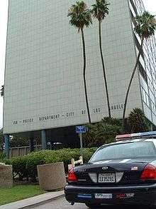 A tall light-colored modernist building with three tall palm trees in front of it and the words "150 – Police Department City of Los Angeles" in capital letters near the bottom. The rear of a parked police car is visible at bottom right