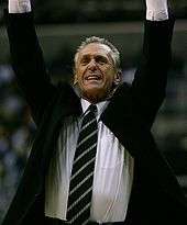 Pat Riley, wearing a black suit with a dark green and silver tie, is throwing his hands in the air as he celebrates a good play.