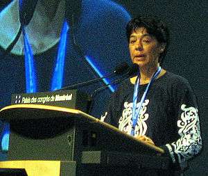 Woman with short dark hair, blue lanyard, and black and white blouse speaking at a podium