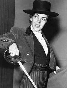 Black and white photograph of a woman wearing a bullfighter's costume, she is holding a sword in one hand and a cape in the other.