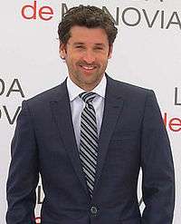 Patrick Dempsey smiling, wearing a navy blue suit.