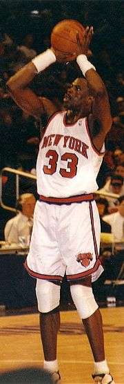 An African American basketball player attempts a jump shot. He wears a white jersey with an orange "NEW YORK" and "33", and spectators can be seen in the background.