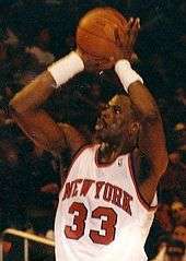 A black basketball player attempts a jump shot. He wears a white jersey with an orange "NEW YORK" and "33", and spectators can be seen in the background.