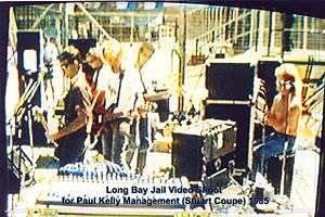 Thirty year old Kelly is shown in left profile playing a guitar in front of a microphone. To his right are two guitarists and a keyboardist amid musical equipment. Behind the keyboard is the drummer at his kit, partly obscured by another microphone. In the background are high fences.