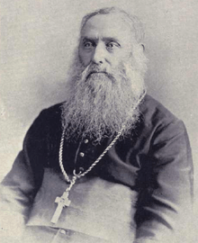 A bearded elderly man wearing a pectoral cross around his neck faces towards the left.