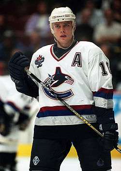 A hockey player stands straight up looking into the distance. He is wearing a white jersey with a red, blue, and silver stripe along the bottom; there are similar stripes on the bottom of his sleeves. The crest of his jersey is of a stylized "C" with a killer whale forming the top part of the logo; his jersey also has a capital "A" patch above his left pectoral.