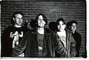 A black and white photograph of five members of the group Pavement standing in front of a brick wall