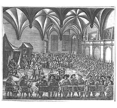 Men gather in a large room, seated on benches around an open center space. Two men read a document to another man seated on a throne.