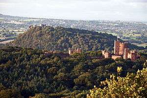 A wooded area with a sandstone castle on the right. Beyond is another wooded hill with the ruins of a castle on its top. In the far distance are fields and houses.