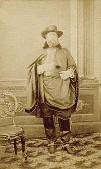 A photographic full-length portrait showing a bearded man wrapped in a short cloak and wearing a wide-brimmed hat and riding boots
