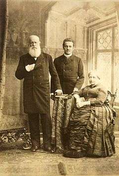 The white-bearded Emperor with his right hand tucked into his formal coat stands next to the right of the seated Empress who wears a hooped gown. In the background stands a younger man in formal dress.