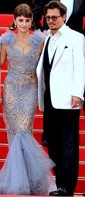 On a staircase with a red carpet stand both a man wearing glasses and a white jacket atop a black business suit, and a woman wearing a blue dress with transparencies.