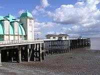 A pier beneath a cloudy sky, with a large white and green building and some smaller huts on it