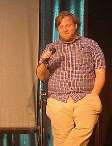 A brown-haired, bearded man in a red button-down shirt and white pants holds a microphone while his other hand rests in his trouser pocket.