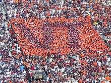 People in the stadium wearing orange and maroon T-shirts to form an image of the letters "VT" in maroon on a square background of orange.
