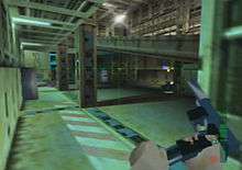 A metallic room with columns and computers located in the farthest side. A hand holding and reloading a gun is seen on the bottom right corner. A crosshair and graphics symbols representing ammunition are also visible.