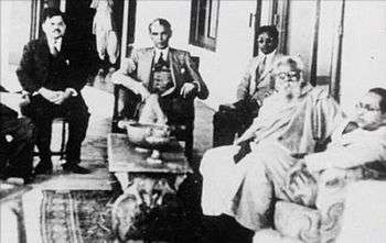 Five men sitting in chairs around a small table. Four of them are wearing suits and one is wearing a shawl and a dhoti. The one wearing the shawl has a flowing white beard.