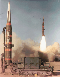 missile erect and prepared for launch while missile is launching in background
