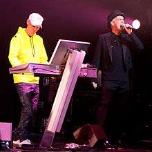 A colour photograph of the two members of the Pet Shop Boys on a stage with a synthesizer and a microphone respectively