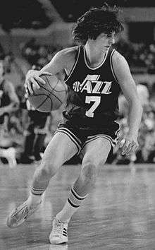 A black and white photo of a man, wearing a basketball uniform that says Jazz with the number 7 below it, is looking to the right.