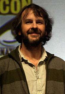 A picture of a bearded man with long black hair. He wears a beige and white striped shirt underneath a charcoal striped jacket.